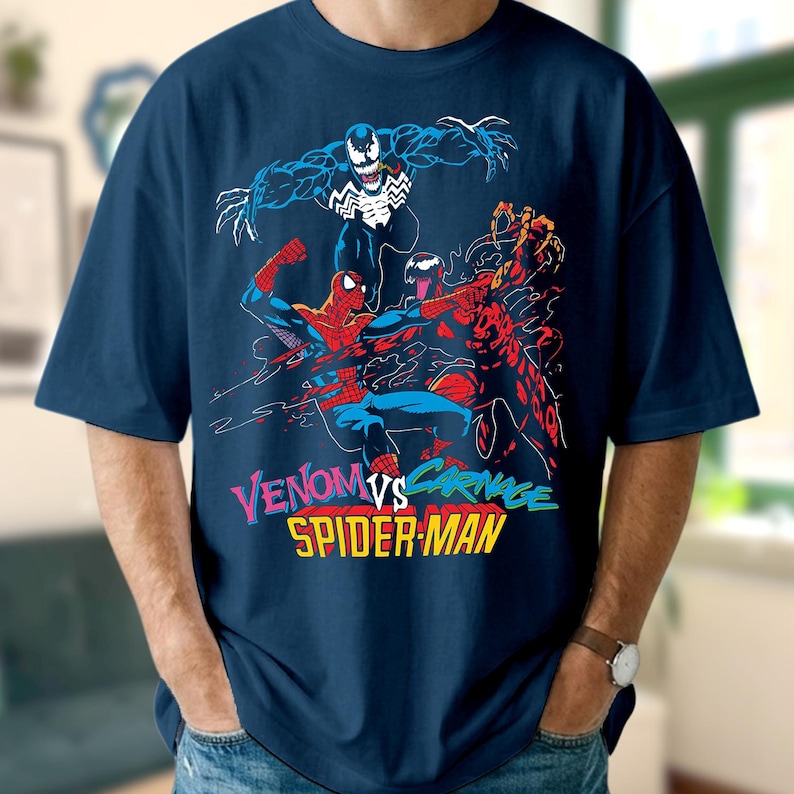 May include: A navy blue t-shirt with a comic book illustration of Venom, Carnage, and Spider-Man. The text "Venom vs. Carnage Spider-Man" is in vibrant colours. The shirt has short sleeves and is made of a soft material.