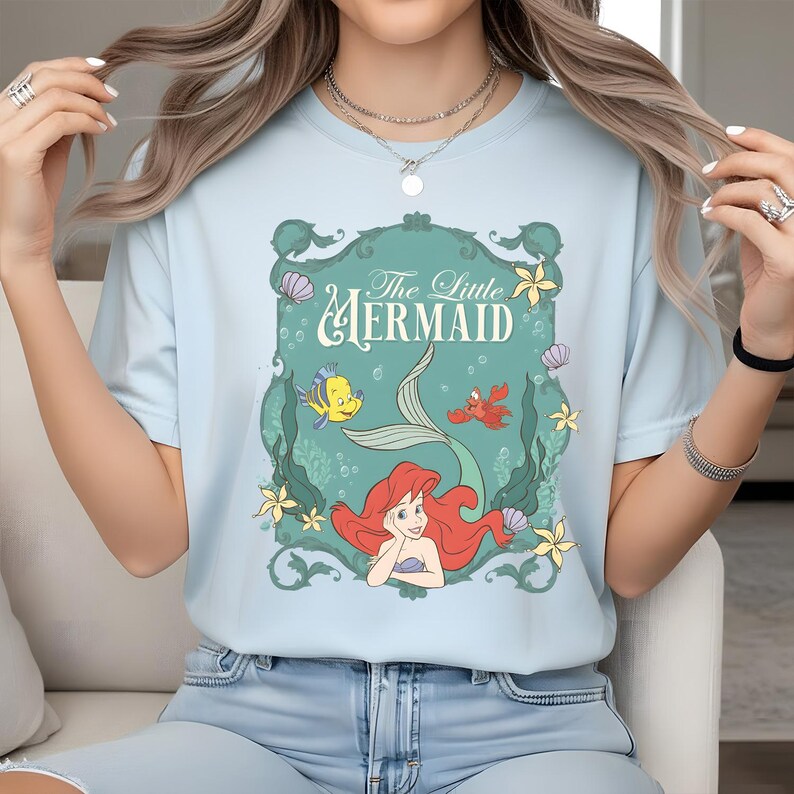 K&ouml;nnte beinhalten: Ein hellblaues T-Shirt mit einem Grafikdruck von Arielle, die Meerjungfrau, mit Fabius und Sebastian in einem dekorativen t&uuml;rkisfarbenen Rahmen. Das Design zeigt gelbe Seesterne, violette Muscheln und gr&uuml;ne Algen auf der Vorderseite.