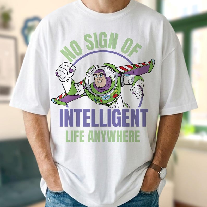 Może przedstawiać: Biała koszulka z kresk&oacute;wkowym wizerunkiem Buzza Astrala i napisem "NO SIGN OF INTELLIGENT LIFE ANYWHERE". Grafika przedstawia Buzza w skafandrze kosmicznym, z zielonymi i fioletowymi akcentami.