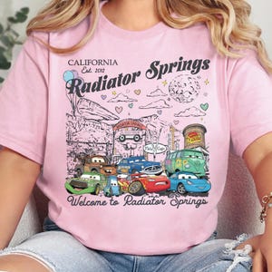 Może przedstawiać: Jasnor&oacute;żowy t-shirt z nadrukiem graficznym przedstawiającym miasto Chłodnica G&oacute;rska z filmu Auta. Wz&oacute;r zawiera r&oacute;żne postacie samochod&oacute;w, znak Chłodnicy G&oacute;rskiej, balony, serca oraz napis California Est. 2012 Radiator Springs.