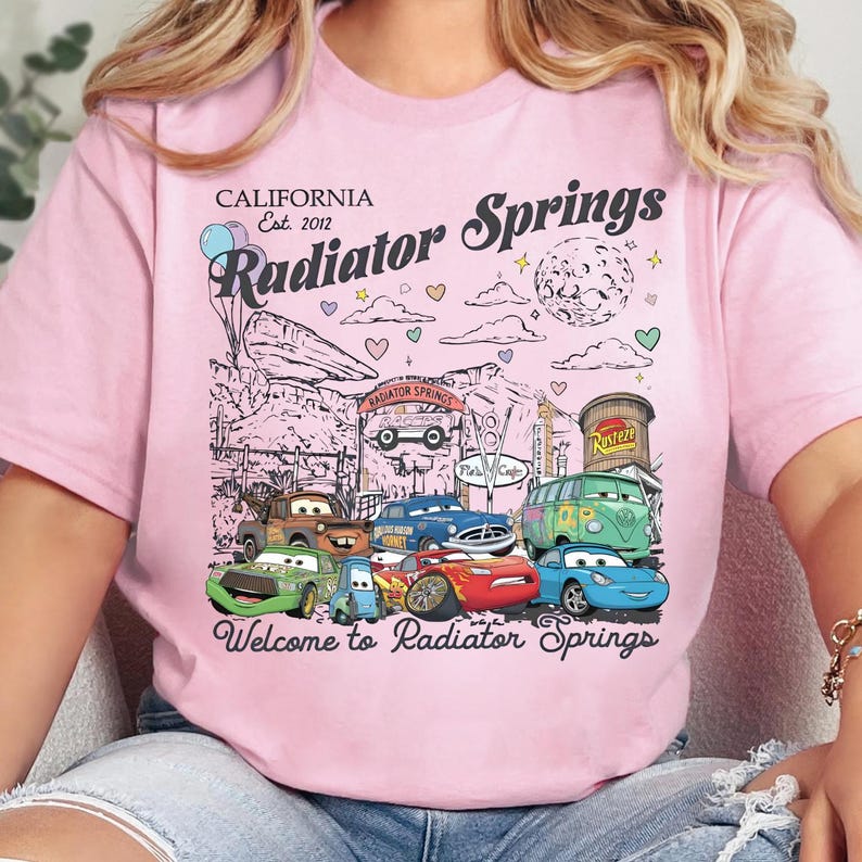 Może przedstawiać: Jasnor&oacute;żowy t-shirt z nadrukiem graficznym przedstawiającym miasto Chłodnica G&oacute;rska z filmu Auta. Wz&oacute;r zawiera r&oacute;żne postacie samochod&oacute;w, znak Chłodnicy G&oacute;rskiej, balony, serca oraz napis California Est. 2012 Radiator Springs.