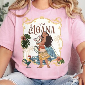 May include: A light pink t-shirt featuring a graphic print of the character Moana holding a piglet, with a rooster standing nearby. The design includes a gold ornate frame, tropical leaves, hibiscus flowers, and the text I AM MOANA.