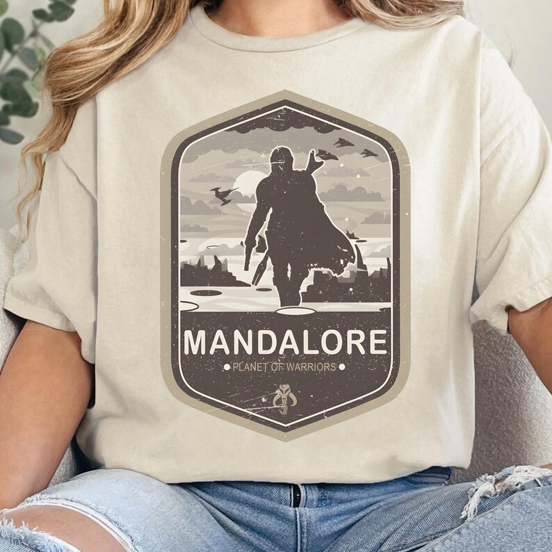 May include: A cream-colored t-shirt featuring a dark brown graphic of a helmeted figure in a cape standing on a landscape. The text reads MANDALORE and PLANET OF WARRIORS, with a small symbol below. The design is enclosed in a hexagonal border.