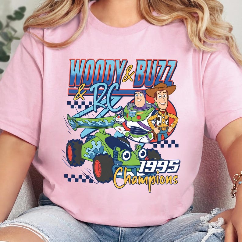 May include: A light pink t-shirt featuring a graphic print of Woody and Buzz Lightyear standing next to a green RC car. The text reads "WOODY & BUZZ & RC" and "1995 Champions" in blue, yellow, and red colours with a black and white checkered pattern.