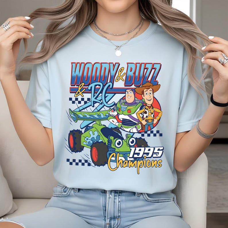 May include: A light blue t-shirt featuring a graphic print of Woody, Buzz Lightyear, and the RC car from Toy Story. The text reads "WOODY & BUZZ & RC" and "1995 Champions" in blue, yellow, and red colours, accompanied by a black and white checkered pattern.