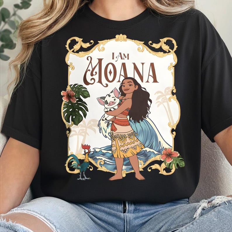 May include: A black t-shirt featuring a graphic print of the character Moana holding a piglet, with a rooster standing nearby. The design includes the text I AM MOANA, framed by a gold ornate border with tropical leaves, hibiscus flowers, and waves.