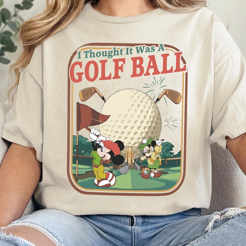 Op de afbeelding: Een cr&egrave;mekleurig t-shirt met een grafische print van twee tekenfilmmuizen die golfen op een groene baan. Het ontwerp bevat een grote golfbal, twee golfclubs en de tekst I Thought It Was A GOLF BALL in rode en groene letters.