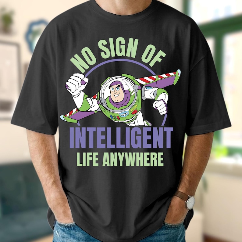 Może przedstawiać: Czarny t-shirt z grafiką Buzza Astrala w locie. Projekt zawiera tekst "NO SIGN OF INTELLIGENT LIFE ANYWHERE" w kolorze zielonym i fioletowym. Koszulka ma okrągły dekolt.