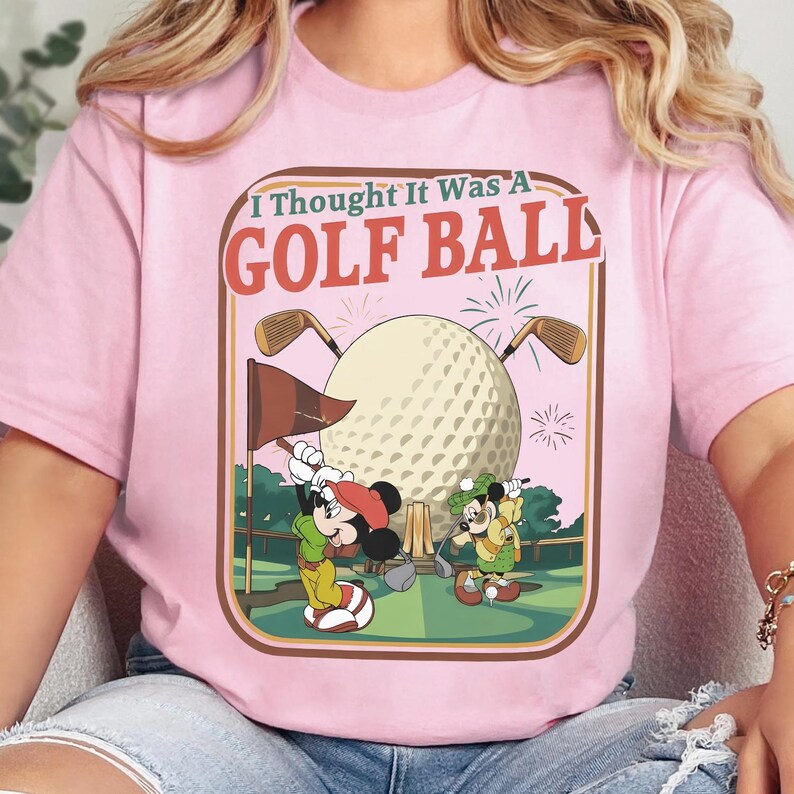 Op de afbeelding: Een lichtroze t-shirt met een grafische print met de tekst I Thought It Was A GOLF BALL. Het ontwerp toont twee stripmuizen op een golfbaan met een grote golfbal, golfclubs, een vlag en vuurwerk in een kader in retrostijl.