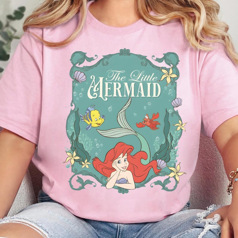 K&ouml;nnte beinhalten: Ein hellrosa T-Shirt mit einem grafischen Aufdruck von Arielle, die Meerjungfrau. Das Design zeigt Arielle, Fabius und Sebastian in einem verzierten blaugr&uuml;nen Rahmen mit Unterwasserelementen wie Seetang, Muscheln und gelben Seesternen.