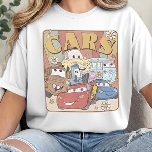 May include: A white t-shirt featuring a graphic print with the text "CARS" in large yellow letters. The design includes illustrations of five cartoon vehicles with faces, surrounded by small star and flower shapes on a muted background.
