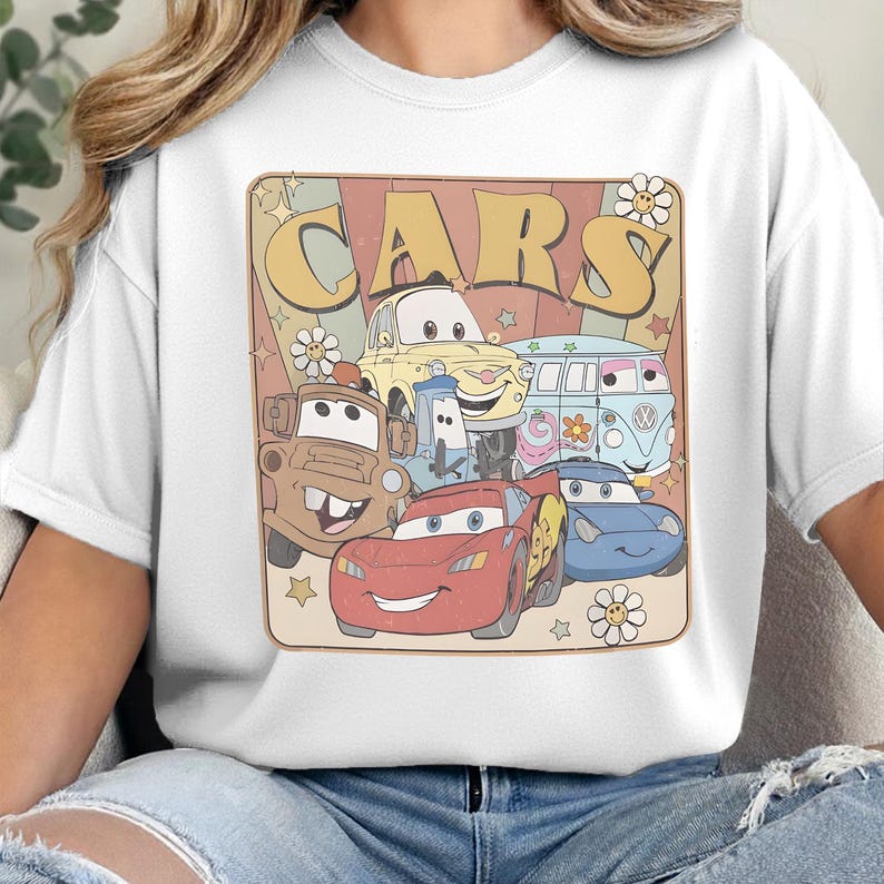May include: A white t-shirt featuring a graphic print with the text "CARS" in large yellow letters. The design includes illustrations of five cartoon vehicles with faces, surrounded by small star and flower shapes on a muted background.