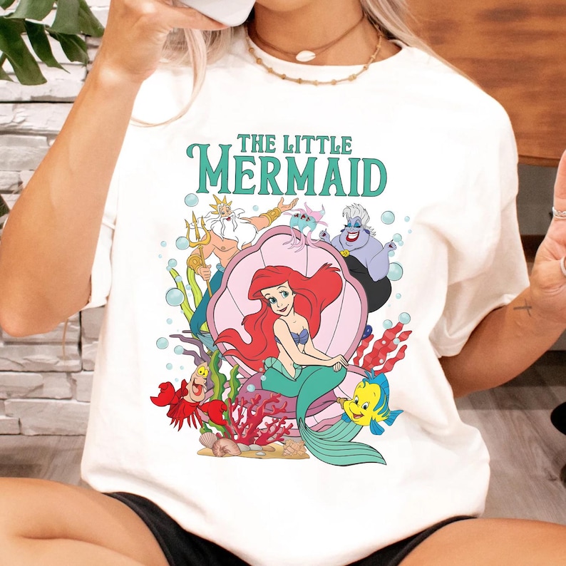 May include: White t-shirt with a vibrant graphic of characters from "The Little Mermaid." The design includes Ariel, King Triton, Ursula, and Flounder, with the title in teal letters. The artwork is set against a backdrop of coral and bubbles.