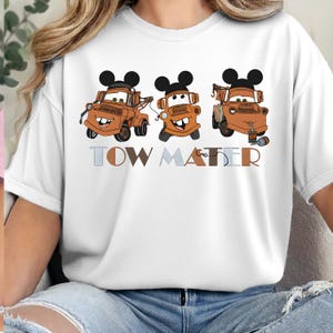 May include: A white short-sleeve t-shirt featuring a graphic of three brown tow trucks, each wearing black mouse ears. Below the trucks, the text TOW MATER is printed in a mix of light blue, brown, and tan block letters with a hook graphic.