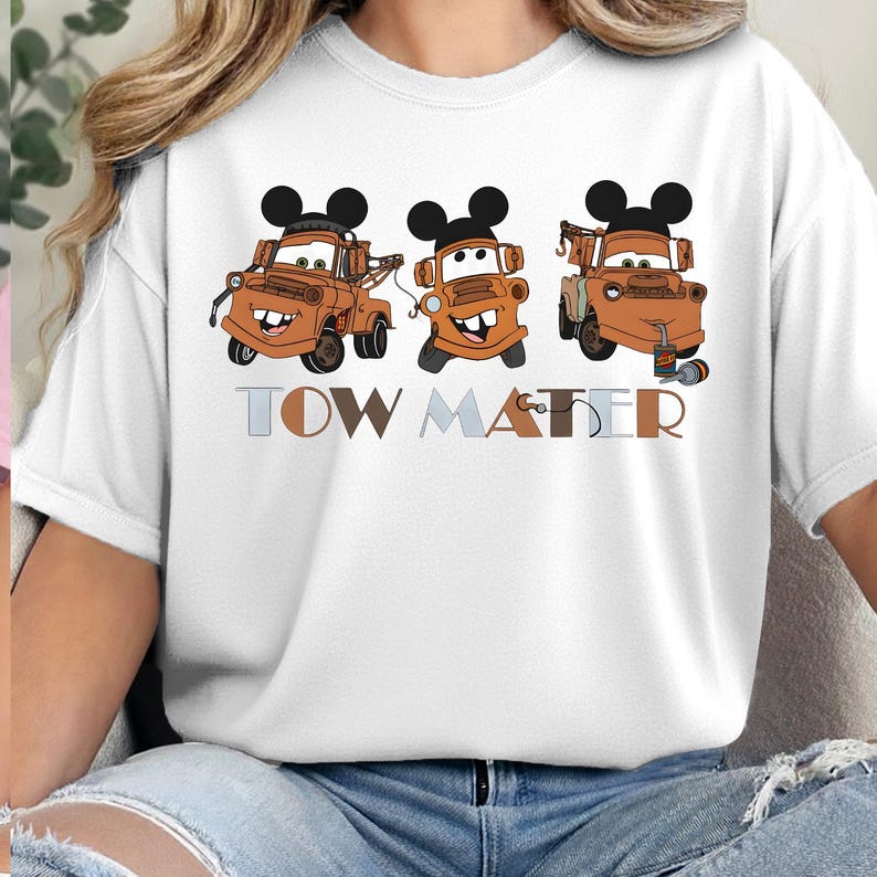 May include: A white short-sleeve t-shirt featuring a graphic of three brown tow trucks, each wearing black mouse ears. Below the trucks, the text TOW MATER is printed in a mix of light blue, brown, and tan block letters with a hook graphic.