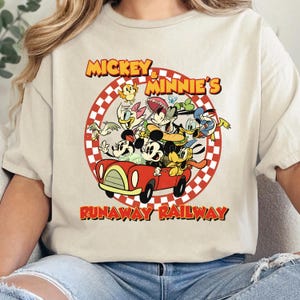 May include: Beige t-shirt featuring a vibrant graphic of Mickey and Minnie Mouse with other cartoon characters in a red car. The text "Mickey & Minnie's Runaway Railway" is displayed in a circular design.