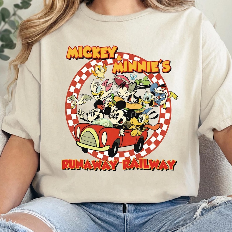 May include: Beige t-shirt featuring a vibrant graphic of Mickey and Minnie Mouse with other cartoon characters in a red car. The text "Mickey & Minnie's Runaway Railway" is displayed in a circular design.