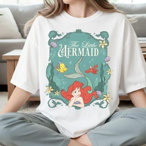 K&ouml;nnte beinhalten: Ein wei&szlig;es T-Shirt mit einem grafischen Aufdruck von Arielle, die Meerjungfrau. Das Design zeigt Arielle, Fabius und Sebastian in einem verzierten t&uuml;rkisfarbenen Rahmen mit Meeresmotiven wie Muscheln, Seetang und gelben Blumen.
