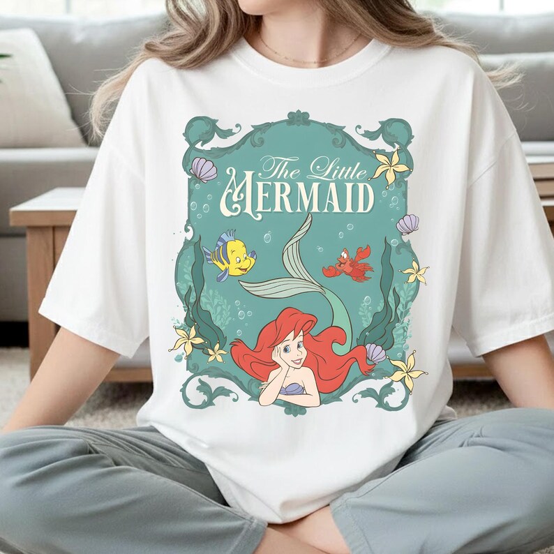 K&ouml;nnte beinhalten: Ein wei&szlig;es T-Shirt mit einem grafischen Aufdruck von Arielle, die Meerjungfrau. Das Design zeigt Arielle, Fabius und Sebastian in einem verzierten t&uuml;rkisfarbenen Rahmen mit Meeresmotiven wie Muscheln, Seetang und gelben Blumen.