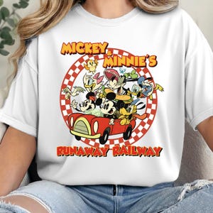 May include: White crew neck t-shirt featuring a graphic of Mickey and Minnie Mouse with other cartoon characters in a red car. The text "Mickey & Minnie's Runaway Railway" is displayed in yellow and red.
