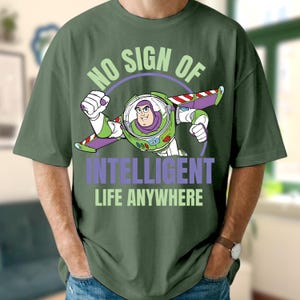 Może przedstawiać: Zielona koszulka z grafiką Buzza Astrala z Toy Story w locie. Tekst "NO SIGN OF INTELLIGENT LIFE ANYWHERE" jest w kolorze zielonym i fioletowym. Grafika to ilustracja z kresk&oacute;wki.