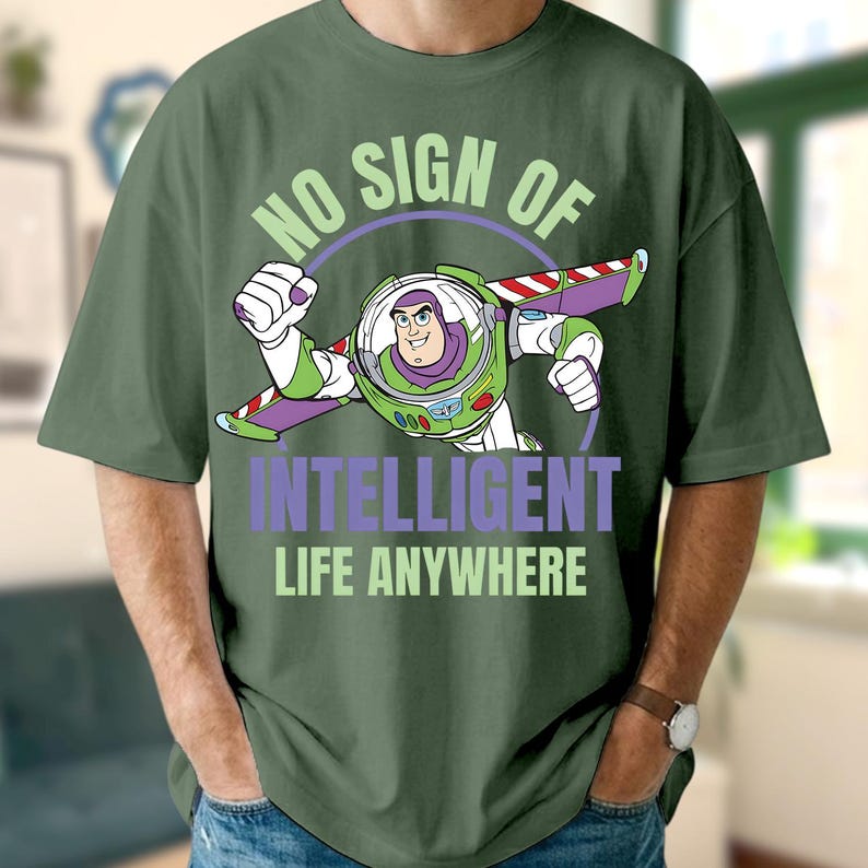 Może przedstawiać: Zielona koszulka z grafiką Buzza Astrala z Toy Story w locie. Tekst "NO SIGN OF INTELLIGENT LIFE ANYWHERE" jest w kolorze zielonym i fioletowym. Grafika to ilustracja z kresk&oacute;wki.