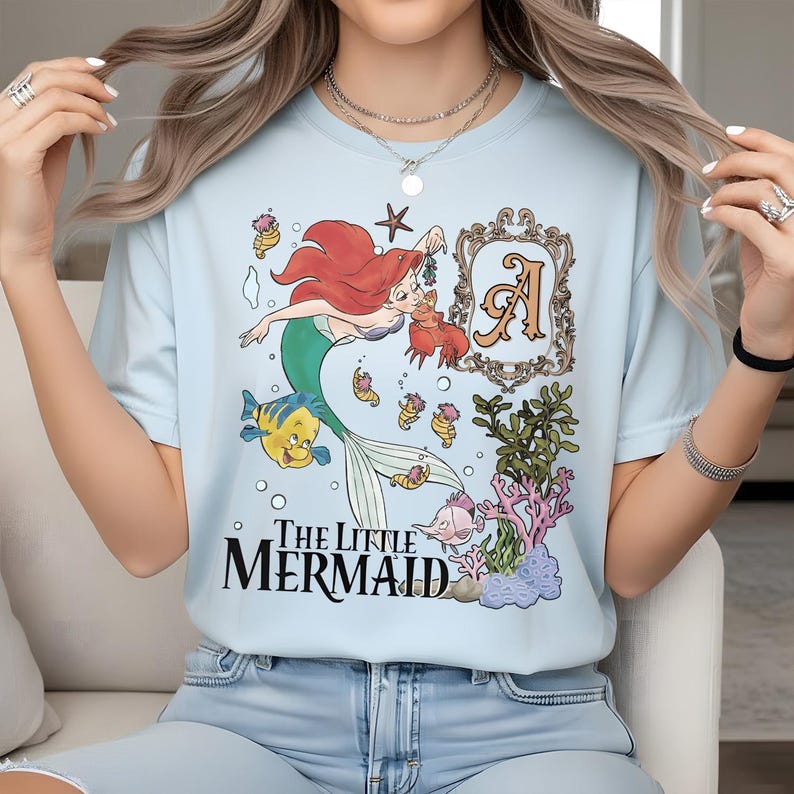 Puede incluir: Una camiseta azul claro con un gr&aacute;fico de Ariel la sirenita, Flounder y Sebasti&aacute;n. El dise&ntilde;o incluye un marco decorativo con la letra A, coral, algas y el texto The Little Mermaid en letras negras en la parte inferior de la prenda.