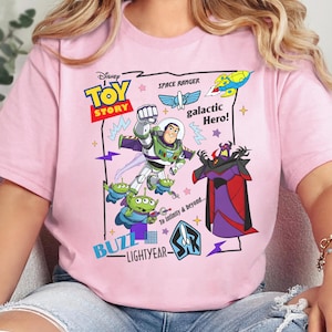 May include: A light pink t-shirt featuring a colorful Disney Toy Story graphic. The design includes Buzz Lightyear, green aliens, and Emperor Zurg, along with text reading Toy Story, Space Ranger, galactic Hero!, and To Infinity & Beyond.