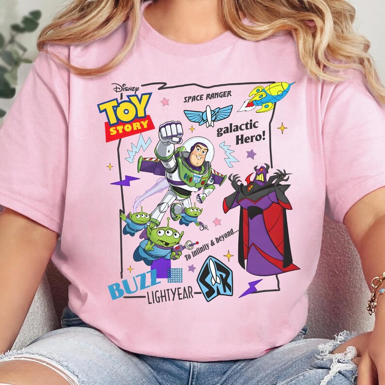 May include: A light pink t-shirt featuring a colorful Disney Toy Story graphic. The design includes Buzz Lightyear, green aliens, and Emperor Zurg, along with text reading Toy Story, Space Ranger, galactic Hero!, and To Infinity & Beyond.
