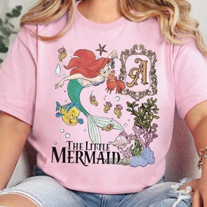 Puede incluir: Una camiseta rosa claro con un gr&aacute;fico de Ariel la sirenita, Flounder y Sebasti&aacute;n. El dise&ntilde;o incluye un marco decorativo con la letra A, varias criaturas marinas, coral y el texto The Little Mermaid en letras de color negro.