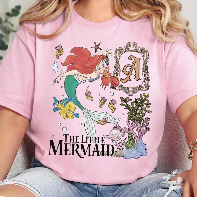 Puede incluir: Una camiseta rosa claro con un gr&aacute;fico de Ariel la sirenita, Flounder y Sebasti&aacute;n. El dise&ntilde;o incluye un marco decorativo con la letra A, varias criaturas marinas, coral y el texto The Little Mermaid en letras de color negro.