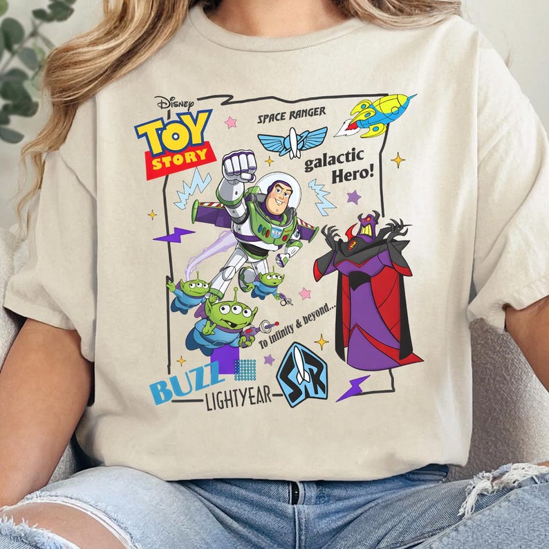 May include: A cream-colored t-shirt featuring a Toy Story graphic with Buzz Lightyear, Zurg, and green aliens. The design includes the text Disney Toy Story, Space Ranger, galactic Hero!, Buzz Lightyear, and To Infinity & beyond... with colorful stars and lightning bolt accents.