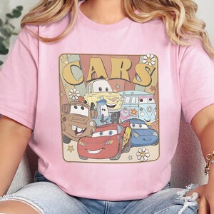 May include: A light pink t-shirt featuring a graphic print with the text "CARS" in large yellow letters. The design includes illustrations of various animated vehicles, including a red race car, a brown tow truck, and a blue van, surrounded by flowers and stars.