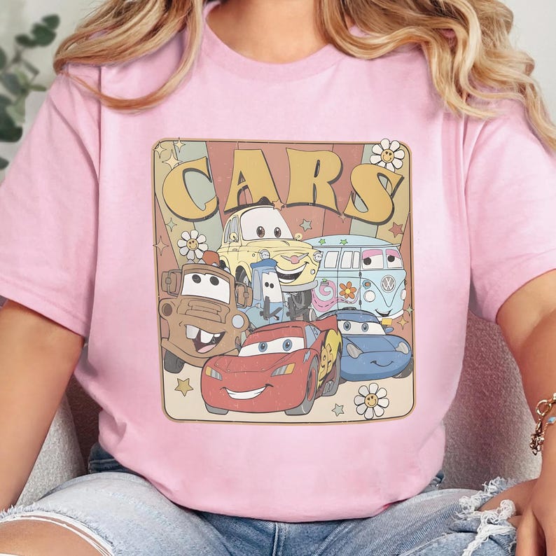 May include: A light pink t-shirt featuring a graphic print with the text "CARS" in large yellow letters. The design includes illustrations of various animated vehicles, including a red race car, a brown tow truck, and a blue van, surrounded by flowers and stars.