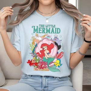 May include: Pale blue t-shirt with a vibrant illustration of characters from "The Little Mermaid." The design includes Ariel, Flounder, Sebastian, King Triton, and Ursula. The words "THE LITTLE MERMAID" are printed above the characters in green.