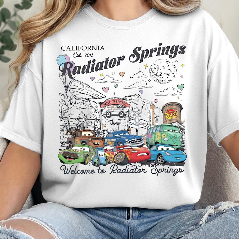 Może przedstawiać: Biały t-shirt z nadrukiem graficznym Radiator Springs oraz tekstem California Est. 2012 Radiator Springs i Welcome to Radiator Springs. Wz&oacute;r zawiera rysunkowe samochody, księżyc, chmury, serca oraz kolorowe balony.