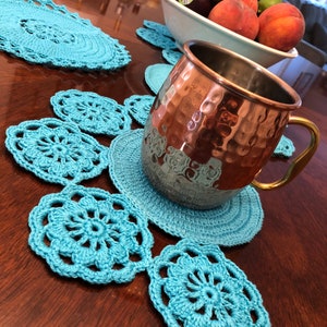 May include: A set of blue crocheted coasters with a hammered copper mug on top. The coasters are circular and have a floral design.