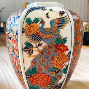 May include: A ceramic vase with a white central panel featuring a blue and orange peacock surrounded by orange and red flowers. The side panels are decorated with an intricate orange and red floral pattern with blue geometric borders.