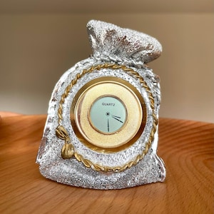 May include: A silver-colored textured bag-shaped desk clock with a gold-toned circular face. The clock features a light green dial with the word QUARTZ printed in black, gold hands, and a decorative gold rope border encircling the clock face.