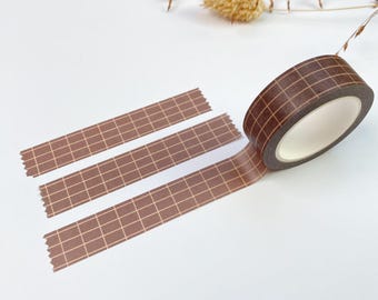 May include: A roll of brown washi tape featuring a white grid pattern. The tape is shown as a single roll with three strips of the same brown and white grid design extended outward, displaying the repeating geometric pattern clearly.