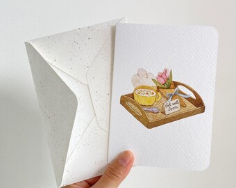 May include: A white greeting card with a watercolor illustration of a wooden tray holding a yellow mug, a bouquet of pink tulips, and a small tag that reads Get well Soon, accompanied by a matching white envelope with dark speckles.