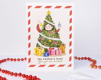 May include: A rectangular greeting card with a red and white striped border featuring an illustration of a person dressed as a Christmas tree holding ornaments. Below the tree are colorful gift boxes and the text You wanted a tree? You got me instead.