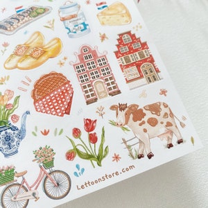 May include: A sheet of stickers featuring illustrations of Dutch culture, including a red brick building, a cow, a bicycle, tulips, and a waffle. The stickers are on a white background and have a whimsical, hand-drawn style.