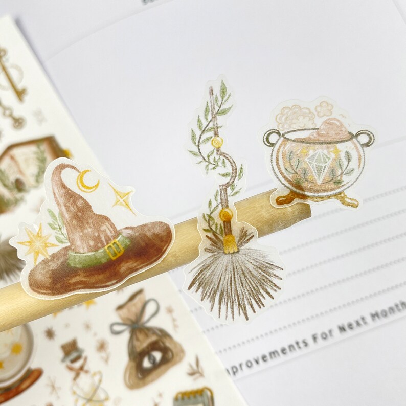 May include: A sheet of clear stickers with a witch's hat, a broom, and a cauldron with a diamond inside. The stickers are on a wooden surface.