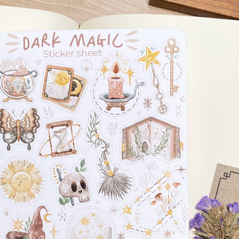 May include: A sticker sheet with a dark magic theme. The sheet features illustrations of a witch's hat, a candle, a book, a butterfly, a sun, a skull, a broom, a key, and a star. The sheet is white with a light brown background.