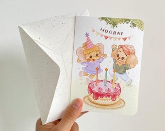 May include: A greeting card with a speckled envelope featuring a watercolor illustration of an elephant and a dog celebrating with a birthday cake. The text HOORAY is printed above them with colorful bunting flags and three lit candles.