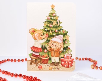May include: A greeting card featuring an illustration of two characters decorating a green Christmas tree with ornaments, lights, and a star topper. One character stands on a wooden stool, while the other holds a string of lights near red gift boxes.