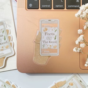 Puede incluir: Una pegatina transparente con una mano sosteniendo un tel&eacute;fono. La pantalla del tel&eacute;fono muestra el texto "Focus on the Good" con ilustraciones florales. La pegatina est&aacute; en una computadora port&aacute;til de color rosa dorado.