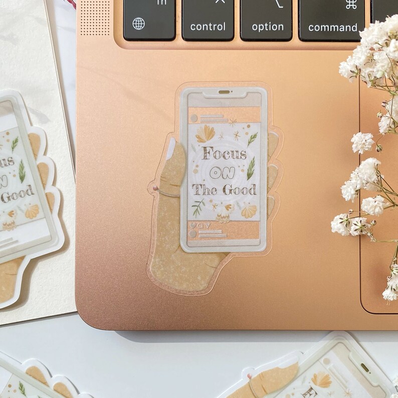 Puede incluir: Una pegatina transparente con una mano sosteniendo un tel&eacute;fono. La pantalla del tel&eacute;fono muestra el texto "Focus on the Good" con ilustraciones florales. La pegatina est&aacute; en una computadora port&aacute;til de color rosa dorado.