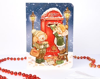 May include: A greeting card featuring a snowy scene with two figures in winter clothing. One figure stands on a stool to place a green envelope into a red post box labeled POST. Two lit street lamps flank the box, and wrapped gifts sit below.